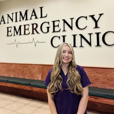 A woman in purple scrubs stands smiling in front of a wall sign that reads "Animal Emergency Clinic" with a heart rate line graphic. There are green benches along the wall behind her.