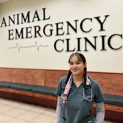 A young woman in scrubs with a stethoscope stands and smiles in front of a wall sign that reads "Animal Emergency Clinic" with a heart rate line graphic underneath.
