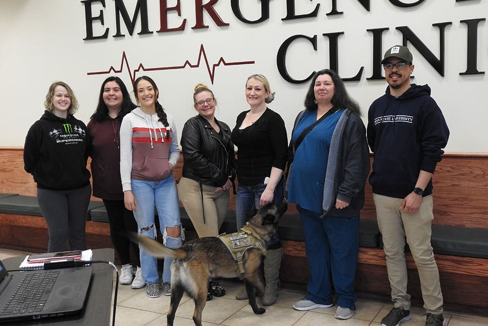A group of six people and a service dog stand in front of a wall with the words "EMERGENCY CLINIC." They are smiling and casually dressed. The service dog, possibly anticipating a visit to the veterinarian, is in the foreground, wearing a vest and looking to the right. They are inside a clinic or hospital lobby.