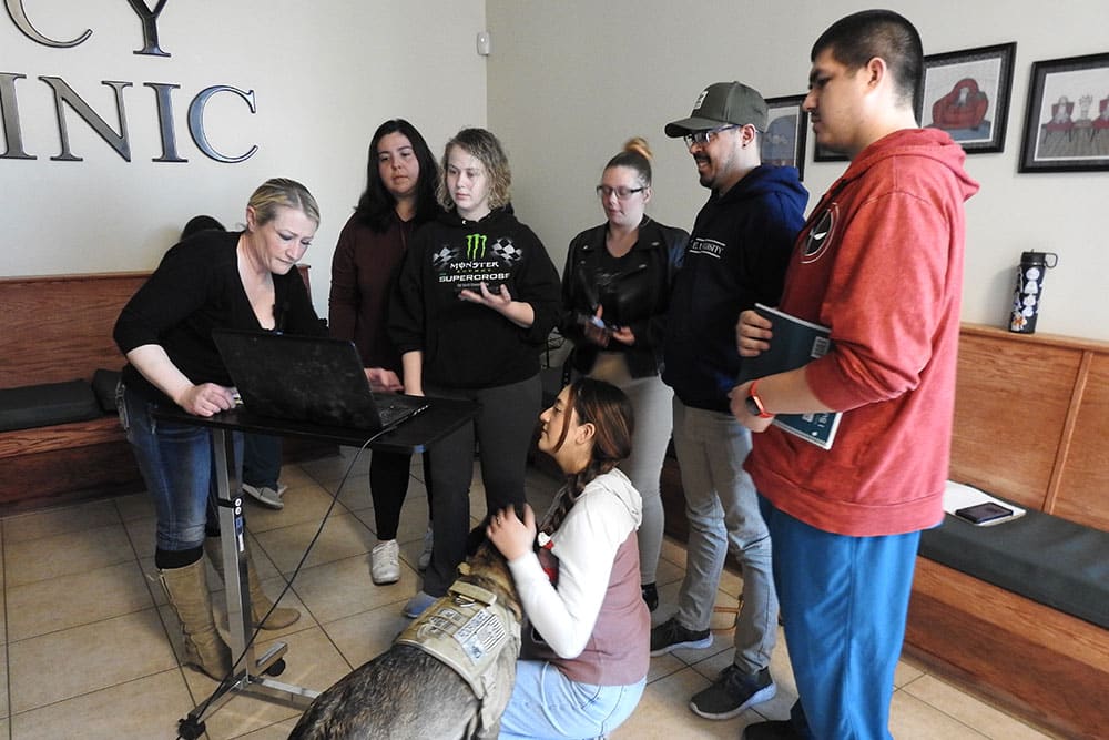 A group of seven people, including one kneeling with a service dog, gather around a laptop on a stand in a clinic waiting room. Operated by a woman standing next to it, the laptop captures their full attention. It appears they might be consulting with the veterinarian via video call.