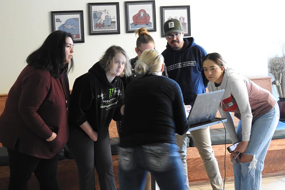 A group of six people, including a veterinarian, gathers around a laptop, intently looking at the screen. The background features framed pictures on the wall. The scene appears to be indoors, with some individuals standing and others bending forward to view the laptop display.