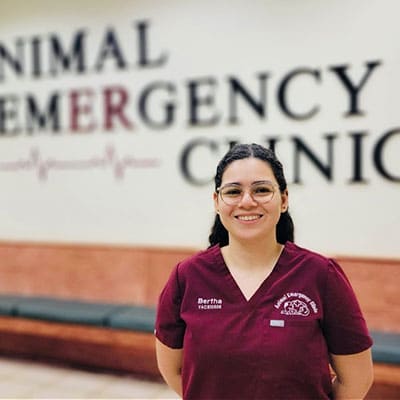 A woman wearing glasses and maroon scrubs stands smiling in front of a wall that reads "Animal Emergency Clinic." The clinic setting appears professional and welcoming.