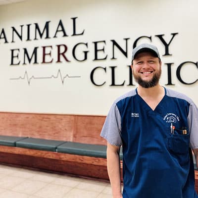 A smiling man in blue scrubs and a cap stands in front of a wall sign that reads "Animal Emergency Clinic" with a heart rate graphic, inside a waiting area with benches.