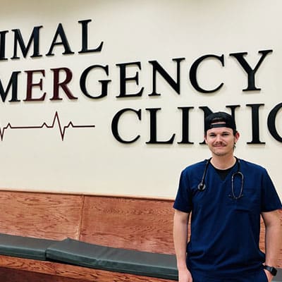 A person in navy blue scrubs with a stethoscope around their neck stands in front of a wall sign that reads "Animal Emergency Clinic" with an EKG line graphic.