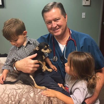 A person in medical scrubs with a stethoscope around their neck, likely a veterinarian, sits with two children and a small black and tan puppy on their lap. The child on the left is looking down at the puppy while the other child on the right reaches out to touch it.