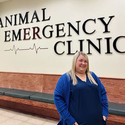A woman with long blonde hair wearing a blue top and cardigan stands smiling in front of a wall sign that reads "Animal Emergency Clinic" with a heartbeat graphic.