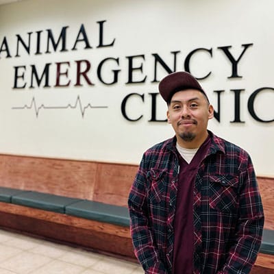 A man in a plaid shirt and cap stands smiling in front of a wall sign that reads "Animal Emergency Clinic" in a waiting area with benches.