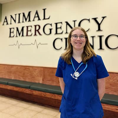 A woman wearing blue scrubs and a stethoscope stands and smiles in front of a wall sign that reads "Animal Emergency Clinic." The setting appears to be a veterinary clinic waiting area.