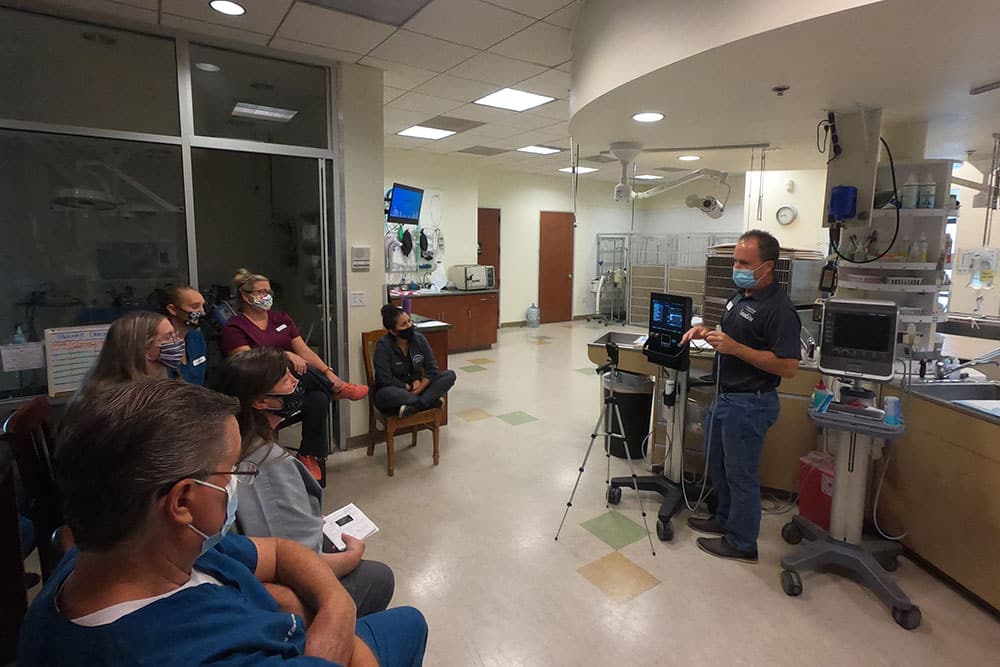 A group of people, wearing masks, sit on chairs in a hospital room, attentively listening to a veterinarian standing and presenting information on a laptop connected to a screen. Medical equipment and monitors are visible in the background.