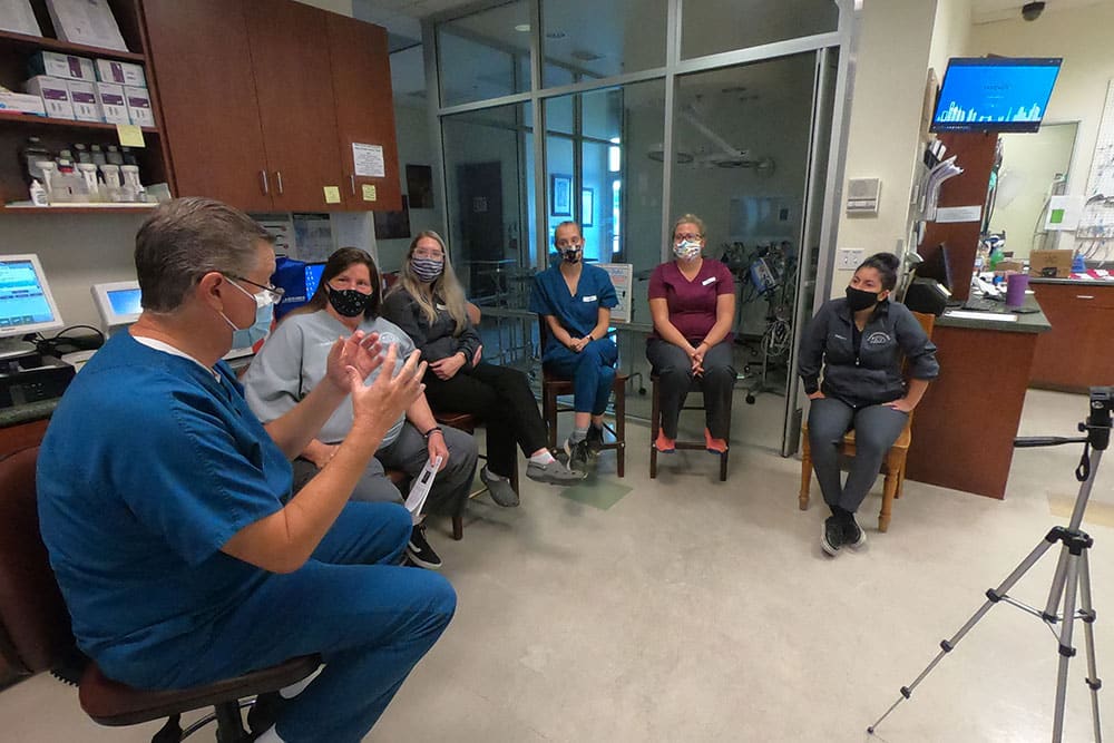 A group of six people, all wearing masks, sit in a semi-circle in a medical office or clinic. A vet in blue scrubs, holding a model, appears to be explaining something to the others who listen attentively. A tripod with a camera is positioned in front of them.