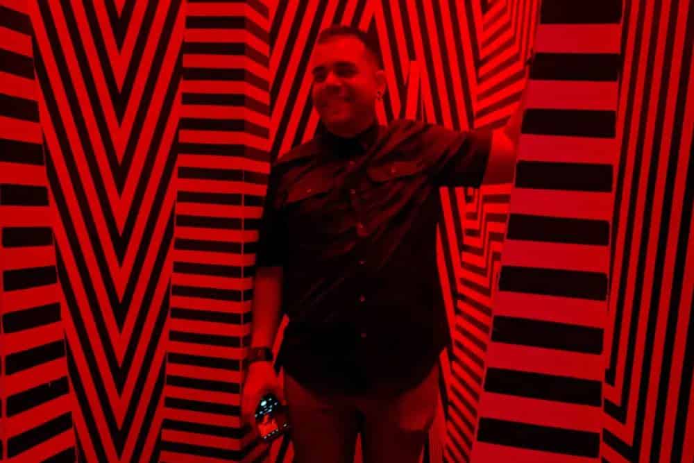 A person smiles while standing in a room with intense red lighting and walls covered with black and white zig-zag and vertical stripes. The veterinarian, holding a camera, wears a dark shirt and pants.