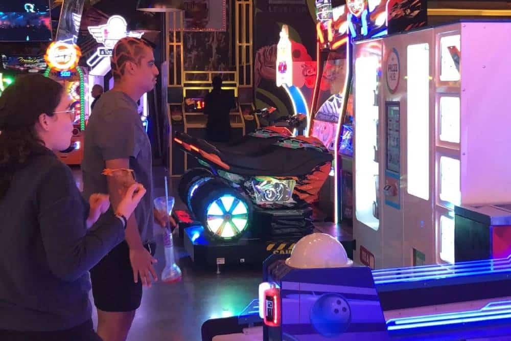 Two individuals are playing arcade games in a brightly lit arcade. The person on the left, a veterinarian unwinding after work, is aiming at a target, while the person on the right observes. In the background, a motorcycle arcade game and neon lights are visible.