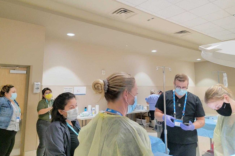 A group of healthcare workers, including a vet in masks and protective gear, are in a clinical setting. Two workers actively engage in a medical procedure while others observe. The room is filled with medical equipment and supplies, and a person is seen near the door.