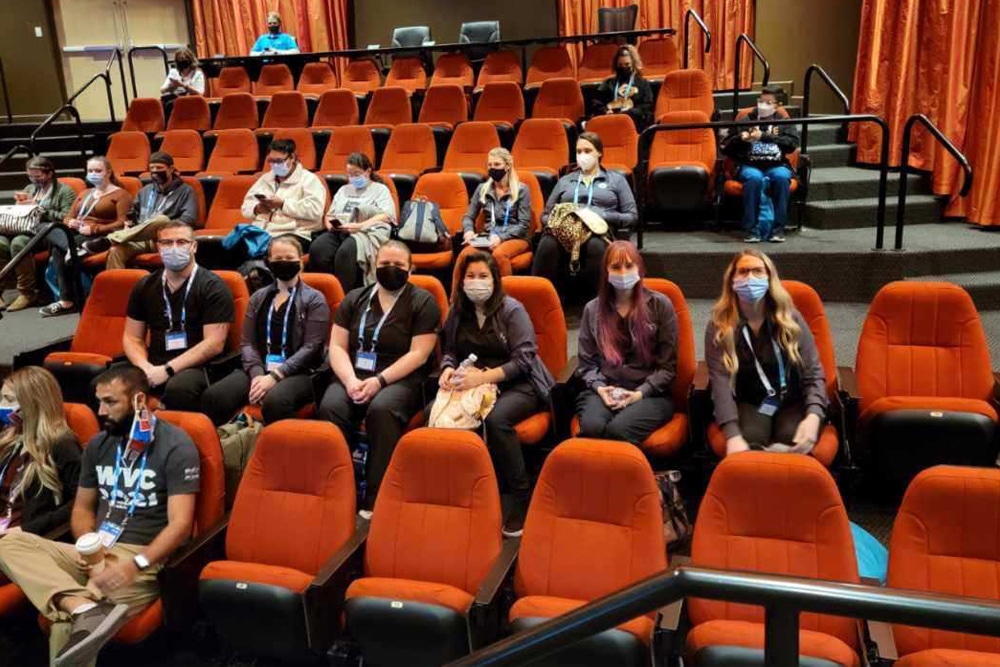 A group of people, including a local veterinarian, all wearing masks, are seated in an auditorium with orange seats. Some individuals are engaged in conversation while others are looking towards the front. The auditorium is partially filled, with several empty seats visible.