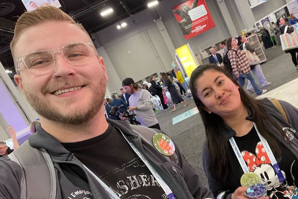 A man with glasses and a beard and a woman with long dark hair are smiling at a busy indoor event. The woman, wearing a Mickey Mouse shirt, is holding a toy. Both have lanyards around their necks. The man, who is clearly a veterinarian, seems to be enjoying the lively atmosphere filled with people and booths in the background.