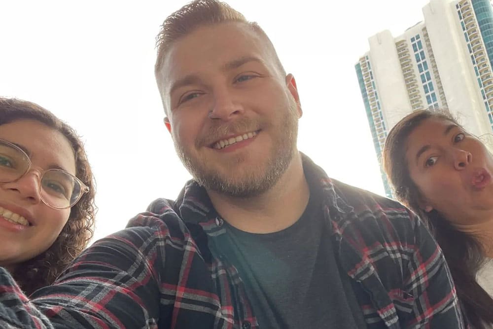 A selfie of three smiling people outdoors with high-rise buildings in the background. The person in the center, a veterinarian, wears a plaid shirt, the person on the left has glasses and curly hair, and the person on the right is making a playful face.
