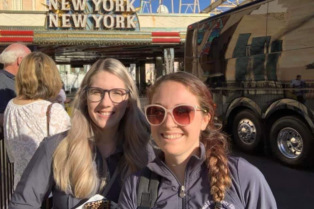 Two smiling women, one a veterinarian, stand in front of the New York New York entrance in what appears to be a city or theme park. Both women wear sunglasses and casual jackets. A bus is parked nearby, and other people are visible in the background.