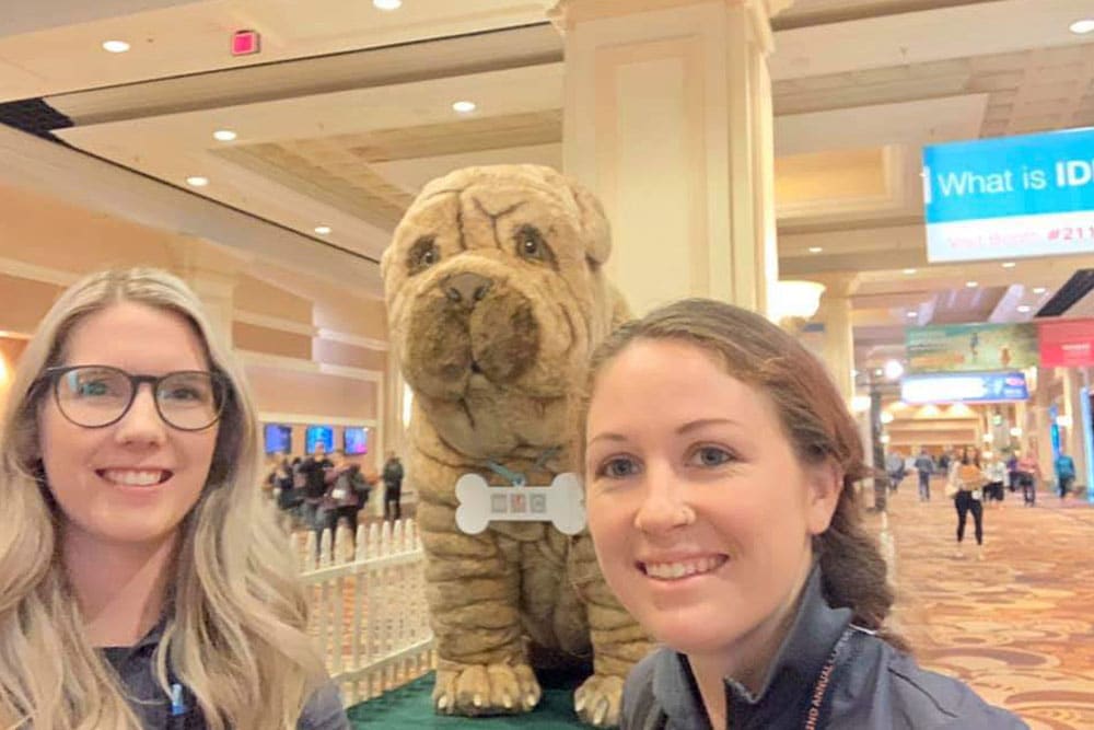 Two women with long hair stand smiling indoors in front of a large plush dog statue wearing a bone-shaped collar. The background reveals a brightly lit indoor event space, possibly organized by a veterinarian, with various signs and people walking around.