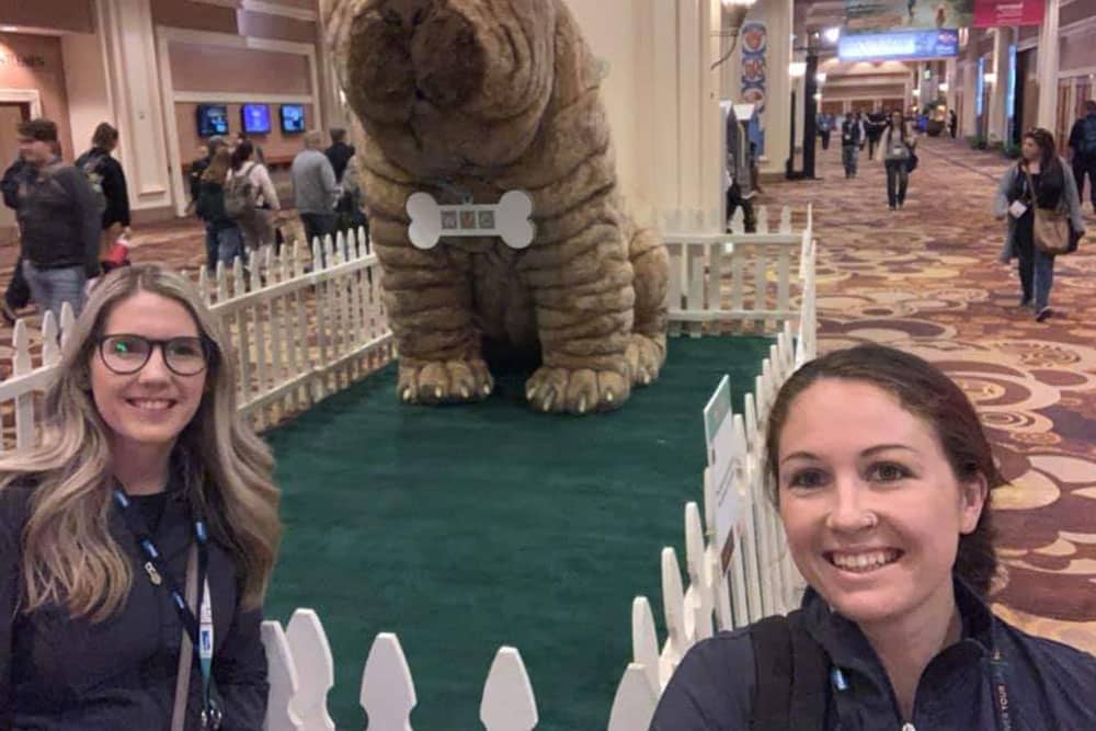 Two women smiling and taking a selfie in front of a large, life-sized dog statue. The statue, enclosed by a small white picket fence, is situated in a spacious, carpeted indoor area. People are walking in the background, perhaps on their way to the nearby veterinarian booth.