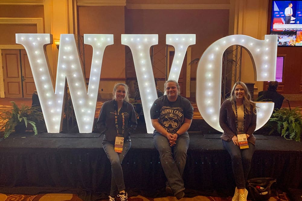 Three people are sitting in front of large, illuminated letters "WVC" at a conference. They are all smiling and appear to be enjoying the veterinary event. The background shows a conference hall decor with lit signs and a few plants. Hanging credentials are visible around their necks.