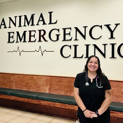 A smiling woman in scrubs with a stethoscope stands in front of a wall sign that reads "Animal Emergency Clinic" with a heart rate graphic underneath the word "emergency.