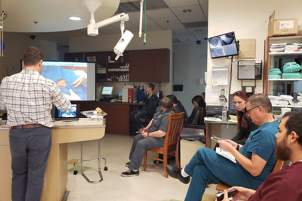 A group of healthcare professionals, wearing scrubs and casual clothing, sit and stand in a medical office while watching a presentation on a projector screen about patient care. Among them is a veterinarian presenting and holding medical equipment. Shelves with medical supplies are in the background.