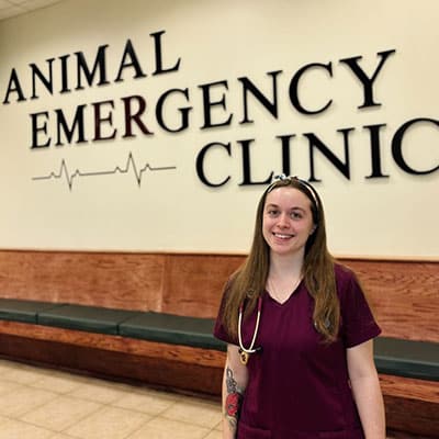 A smiling woman in maroon scrubs with a stethoscope stands in front of a wall sign that reads "Animal Emergency Clinic." Benches line the wall behind her.