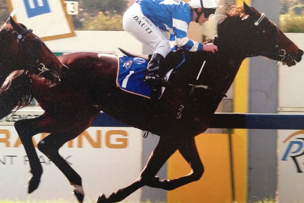 A jockey, dressed in a blue and white striped uniform, rides a dark brown racehorse during a race. The horse is mid-stride, and both horse and rider appear focused on the track ahead. A veterinarian stands by near the advertising signs visible in the background.