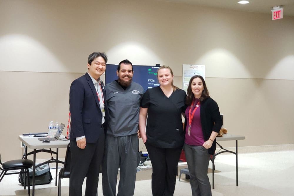 Four people, including a veterinarian, are standing together in a well-lit room with smiles, posing for a group photo. Behind them is a table with various items on it and a screen displaying indistinct content. The people are casually dressed, and the setting appears to be informal and friendly.