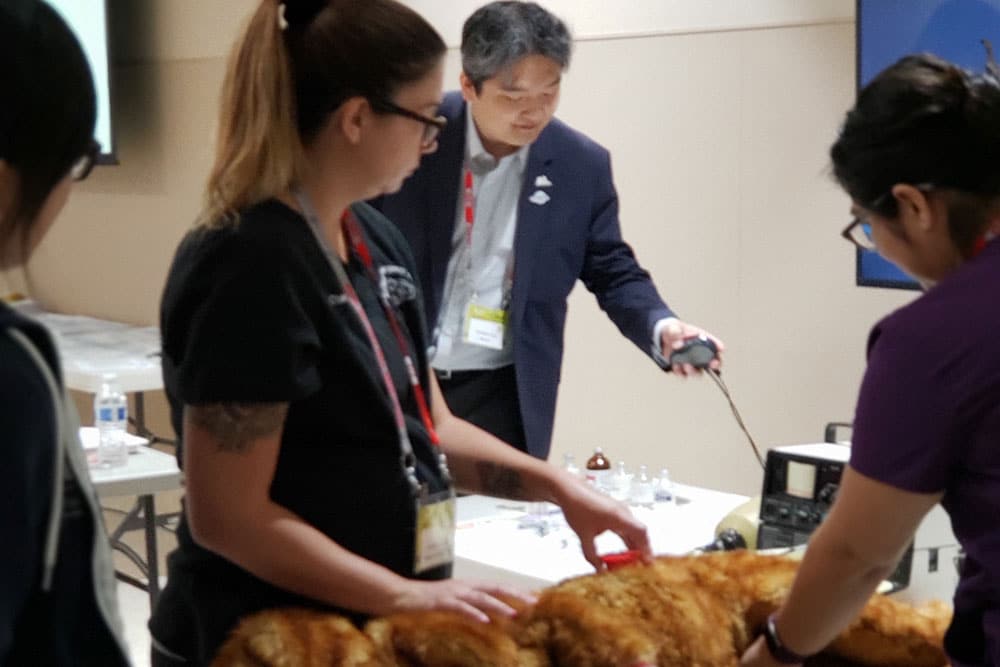 A group of people stand around a table with a large, furry object in the center. One person, possibly a veterinarian, is using a device on the object while another looks on. In the background, a man in a suit holds an instrument and observes the scene. The table has various small items.