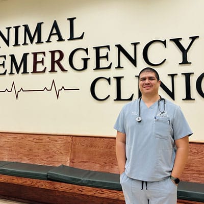 A veterinarian in light blue scrubs stands in front of a wall with the words "Animal Emergency Clinic" and a heartbeat graphic. He has a stethoscope around his neck and is smiling.