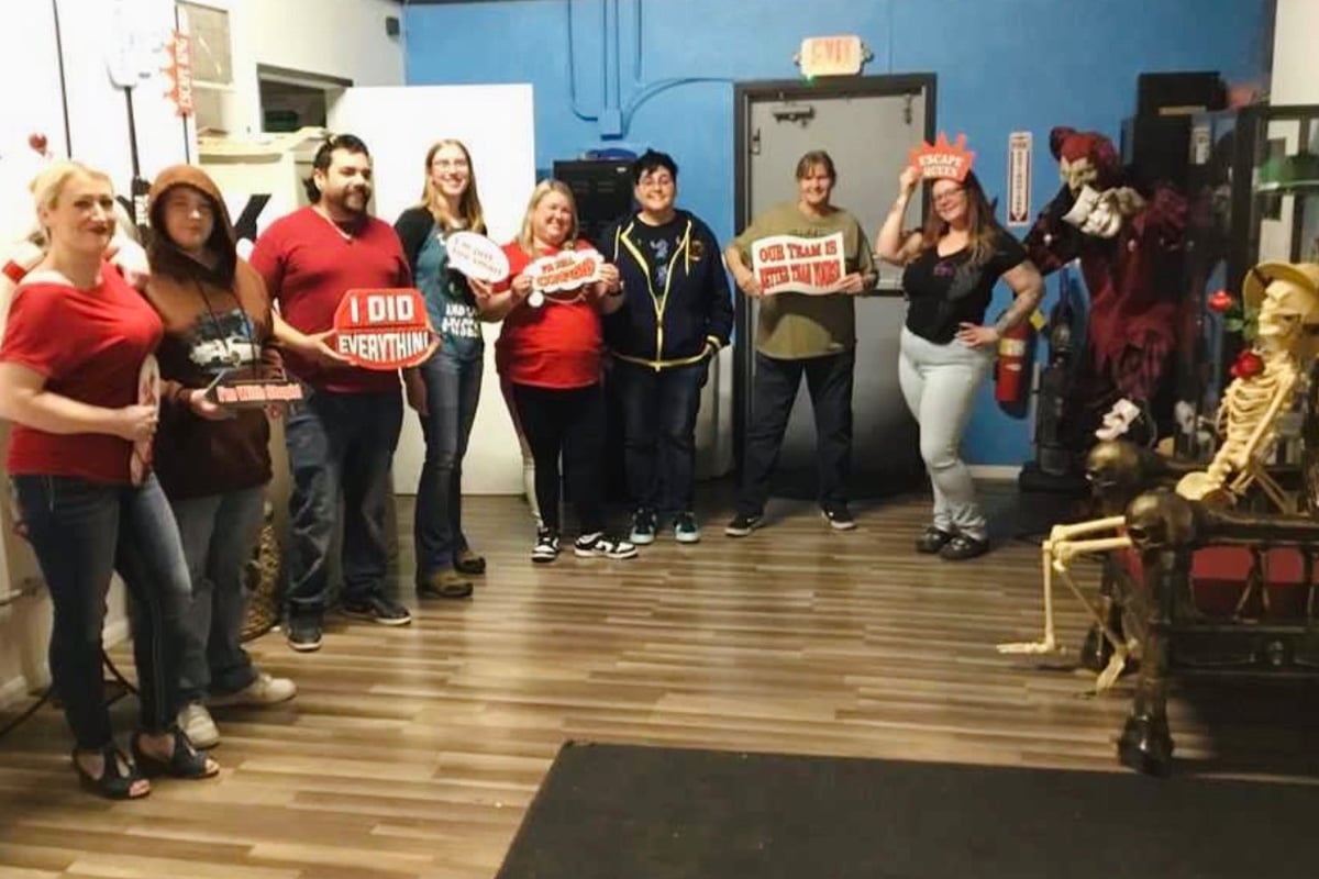 staff-event-16 A group of eight people stand in a room holding playful signs. Some are smiling, and the room is decorated with Halloween props including skeletons and a creepy mannequin. The floor is wood, and the walls are blue and white.