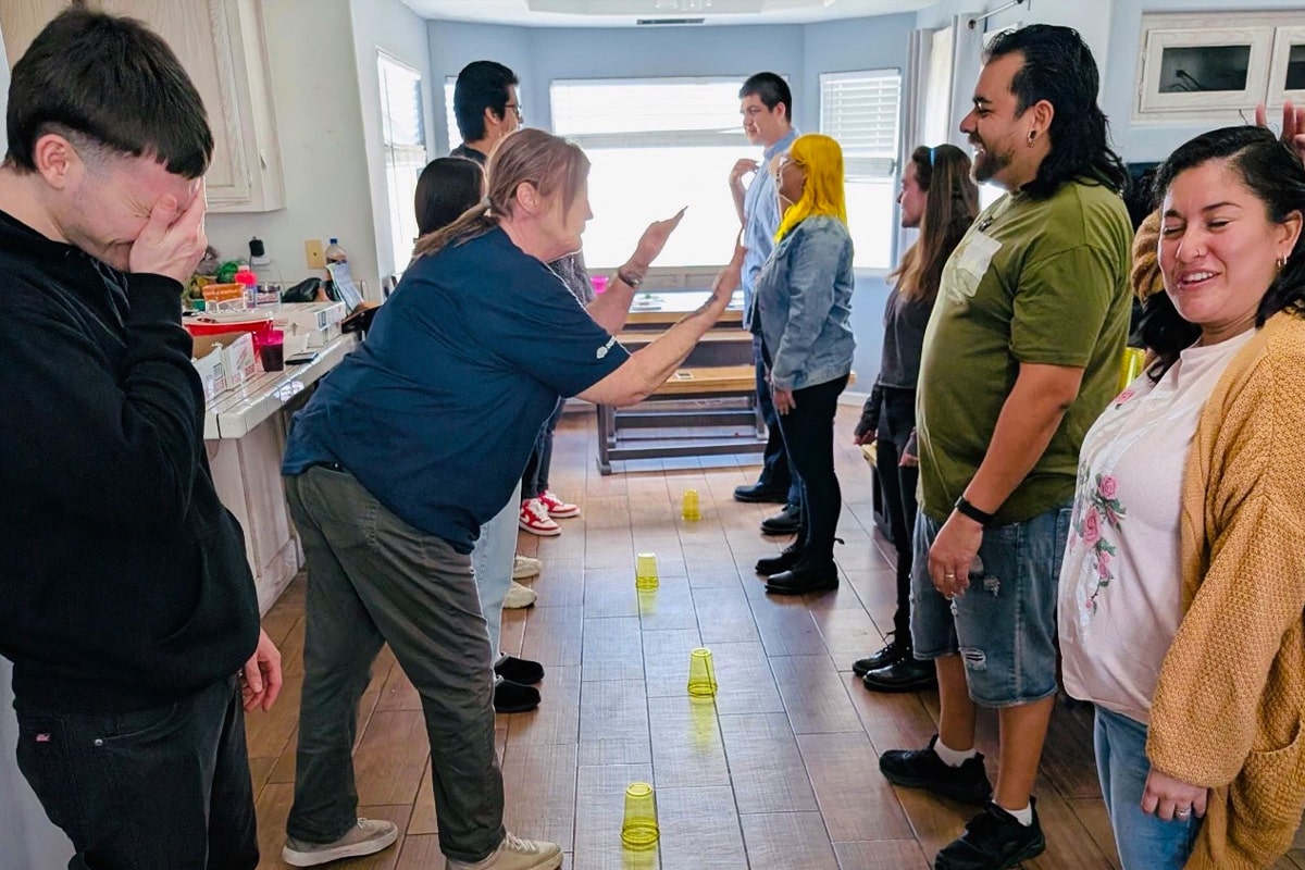 staff-event-4 A group of people stand facing each other in two lines inside a kitchen, laughing and playing a game with yellow plastic cups arranged on the floor between them. The atmosphere is cheerful and lively.