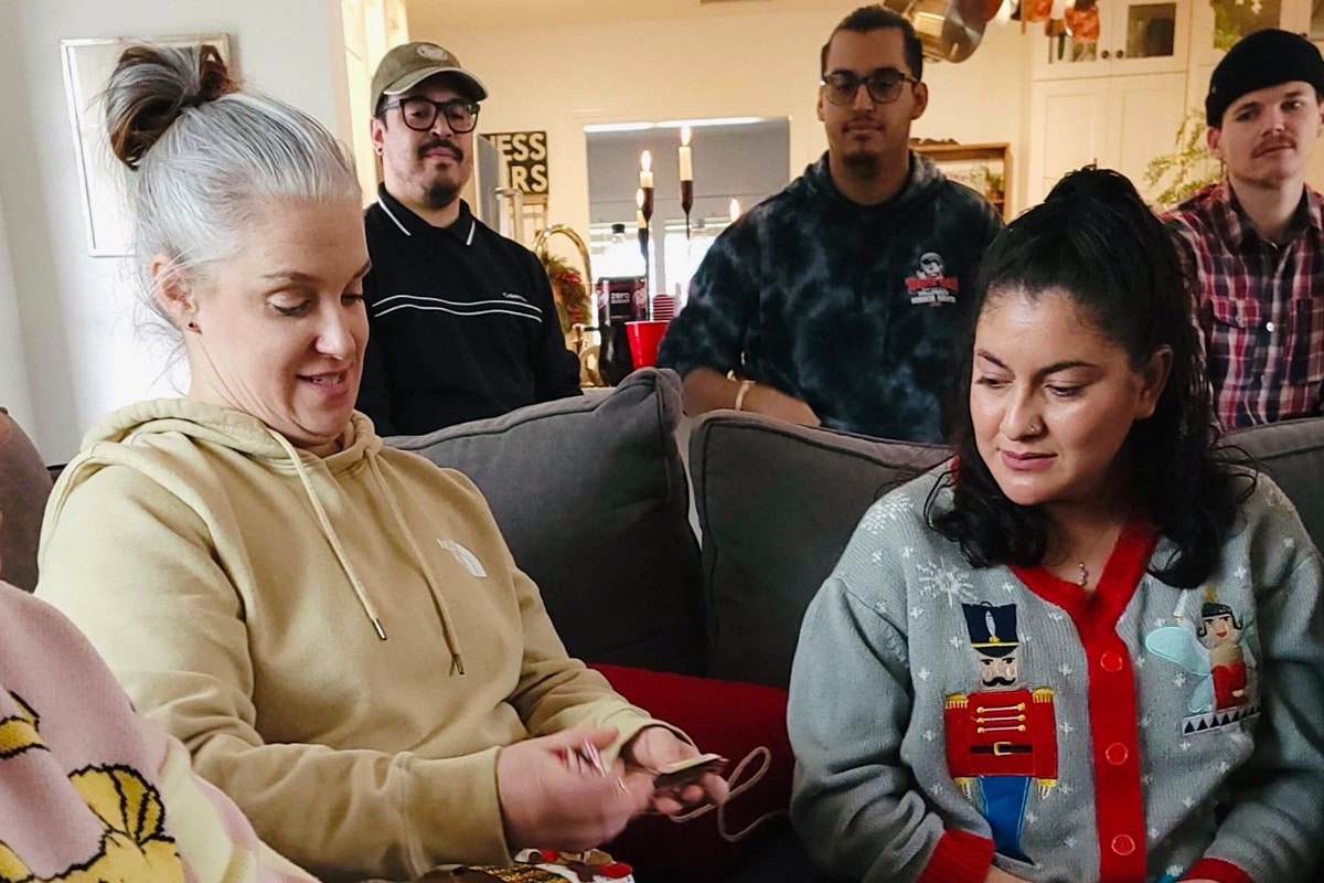 staff-event-9 A group of five adults sit together indoors. Two women sit on a couch in the foreground, one wearing a beige hoodie and the other a festive sweater, while three men sit and stand behind them, all engaged and watching the women.